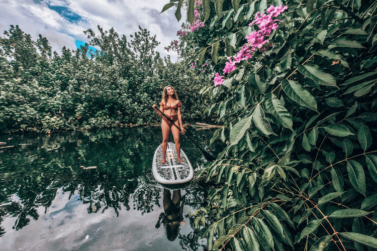 Woman paddleboarding down river with lush plants and flowers